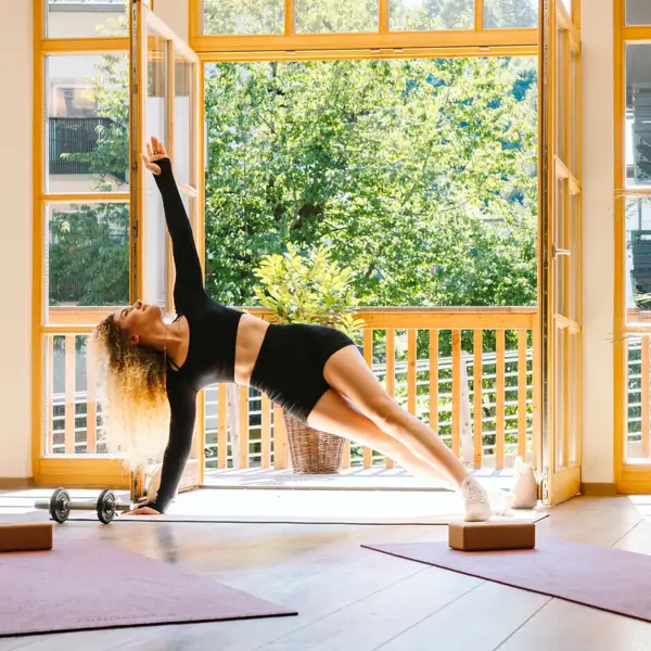 A woman does a side plank in a room with windows.