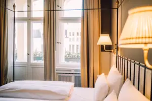 Gemütliches Hotelzimmer im HENRI Berlin mit hohen Decken, hellem Fenster und eleganten Nachttischlampen im Gründerzeit-Stil.