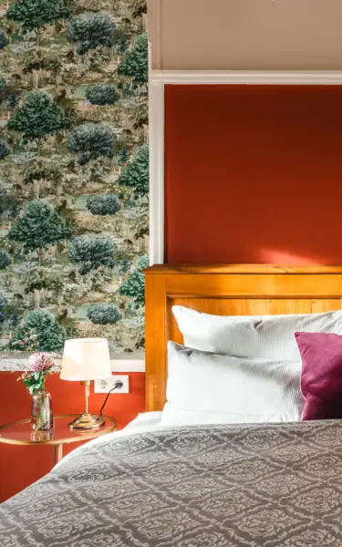 A cozy indoor scene with two wooden chairs draped in soft white sheepskin facing a sunlit window. A small round table with a glass vase holding flowers stands between the chairs, with sunlight streaming through the window.