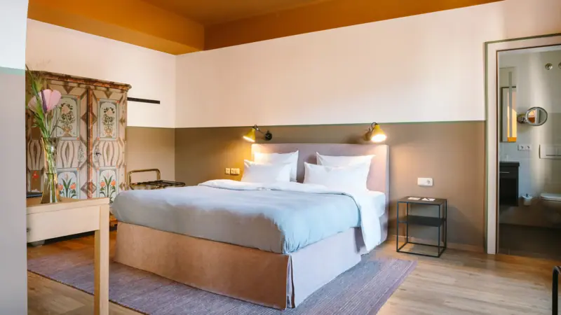 A hotel room at the HENRI Garmisch-Partenkirchen with a large cosy bed, an old wardrobe and a door to the adjoining bathroom.