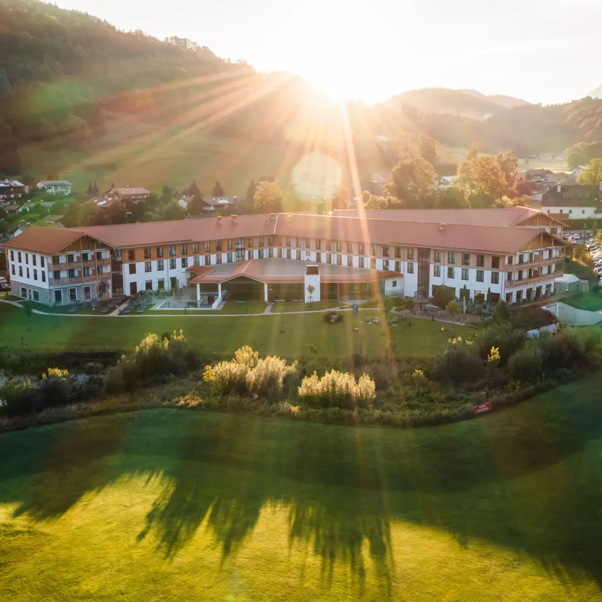 Aerial view of a large hotel with a spacious garden in the Alpine landscape, sunbeams shine over the hills and the morning sky