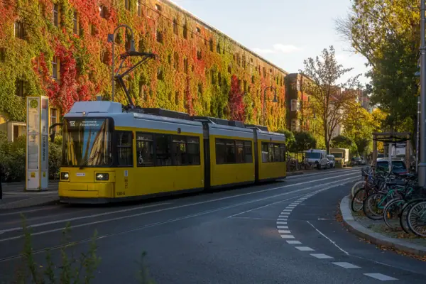 Berlin Lifestyle Tram Autumn A yellow Berlin tram at a stop in front of an autumnal red and green house facade under a blue sky.