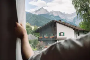 A hand is holding a curtain in front of a window, with mountains in the background.