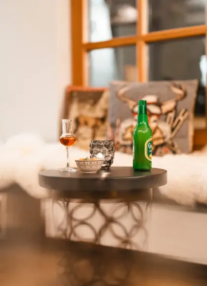 Cozy evening setup featuring a small round table with a glass of liqueur, a green beer bottle, a decorative candle holder, and a bowl of snacks.