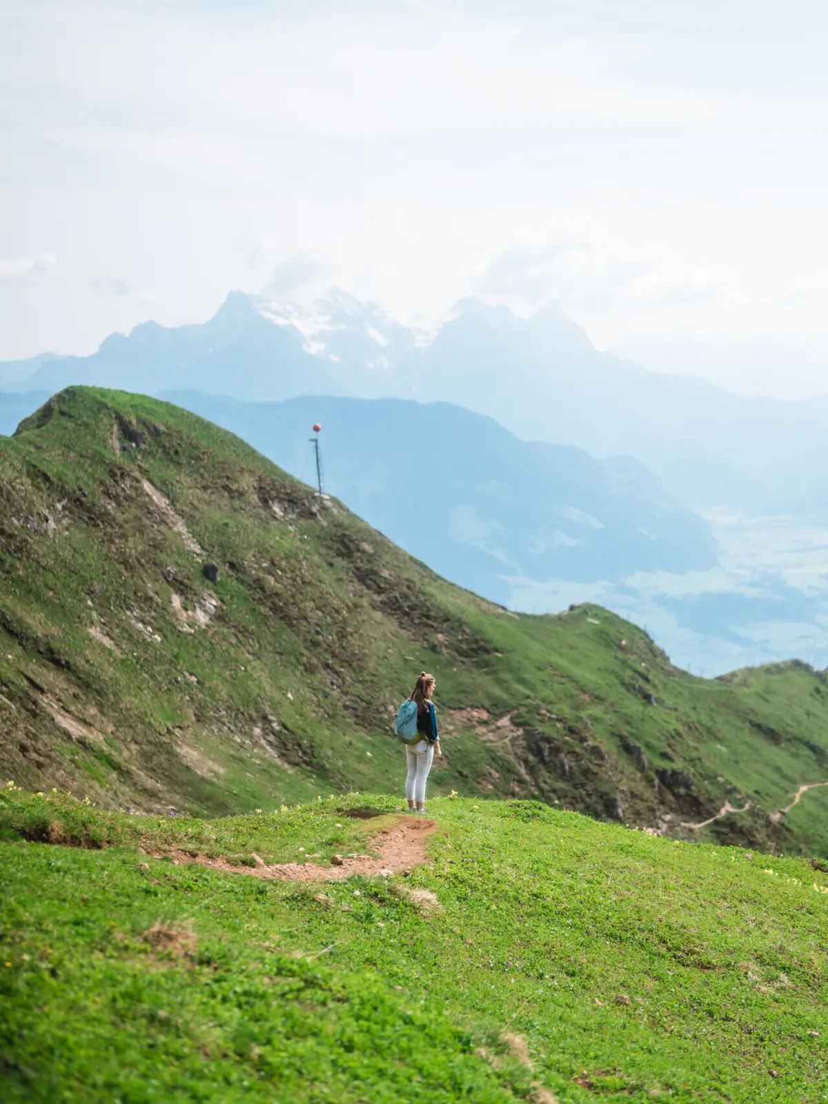 A woman stands on a path in a grassy area with mountains in the background.