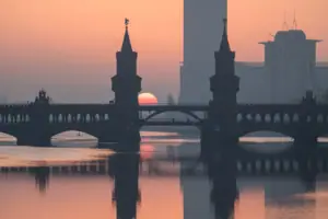 Silhouette of the Oberbaumbrücke in Berlin at sunrise, with the towers and the reddish sun reflected in the calm waters of the Spree.