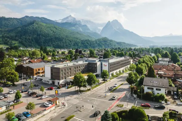 The aja Garmisch-Partenkirchen with many buildings and trees under a cloudy sky and mountains in the background.