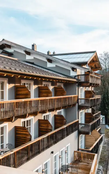 A picturesque winter village nestled in a valley surrounded by snow-covered mountains. Charming alpine-style buildings with snow-dusted roofs are scattered amidst trees, with majestic peaks rising in the background under a bright blue sky.