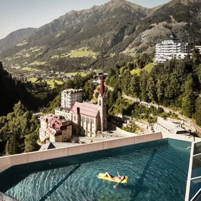 A person swims with a yellow air mattress in a rooftop infinity pool on a tall building with mountains in the background.