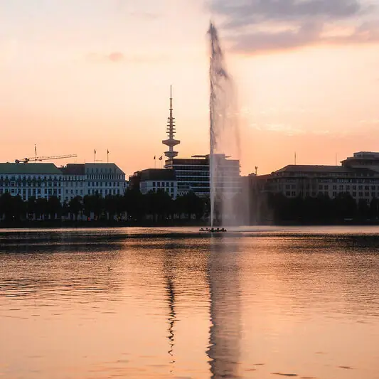 Fountain in a body of water with buildings in the background.
