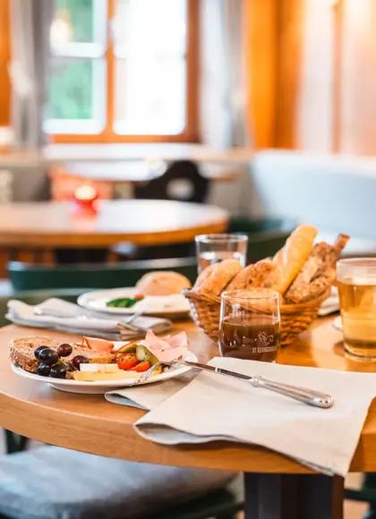 Cozy dining setup in a rustic restaurant with a wooden table set for breakfast, featuring plates of cold cuts, cheese, bread, and fresh vegetables.