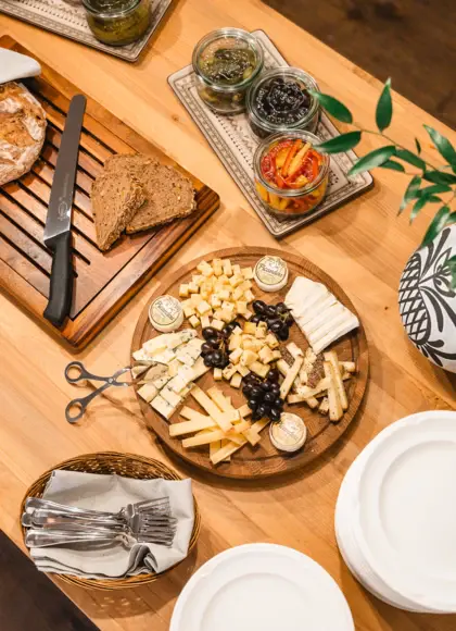 Wooden table with a cheese platter featuring a variety of cheesesand a stack of white plates with cutlery nearby.