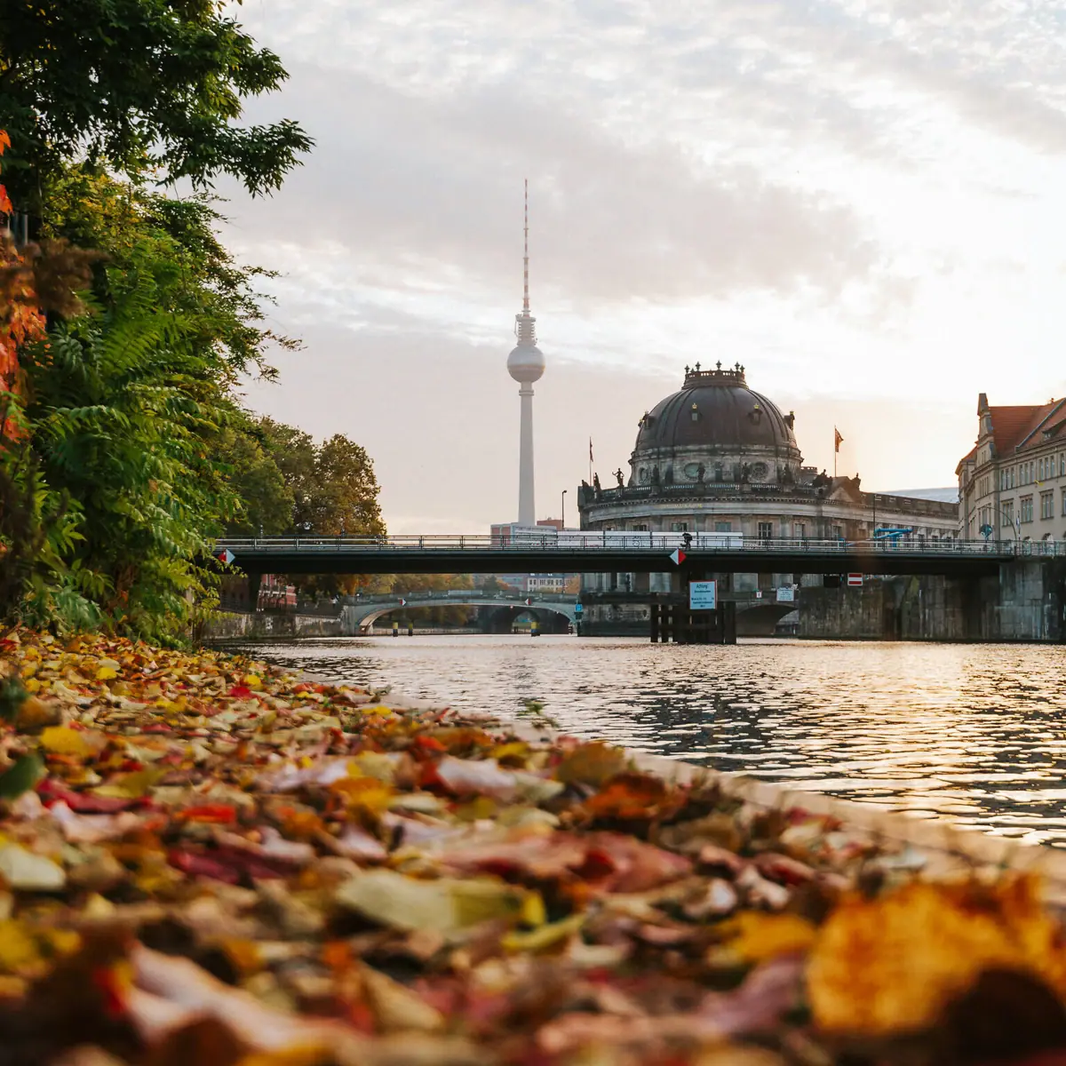 The River Spree with a bridge and the Berlin TV Tower in the background, surrounded by autumnal trees and clouds in the sky.