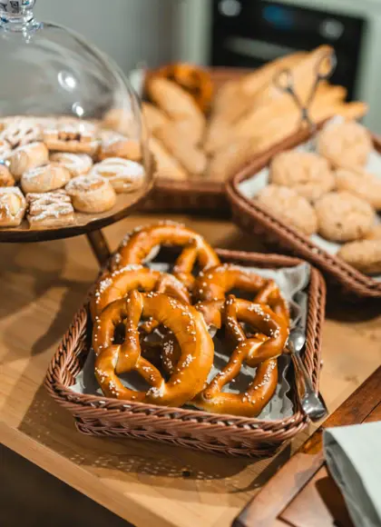 Rustic breakfast setup featuring freshly baked pretzels with coarse salt in a wicker basket, accompanied by other baked goods such as rolls, baguettes, and pastries under a glass dome, arranged on a wooden table.