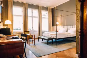 Spacious, light-flooded hotel room at the HENRI Berlin with a classic four-poster bed, herringbone parquet flooring and stylish vintage furniture in front of high windows.