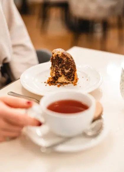 Close-up of a slice of marble cake served on a white plate alongside a cup of tea in a white porcelain cup held by a hand. 