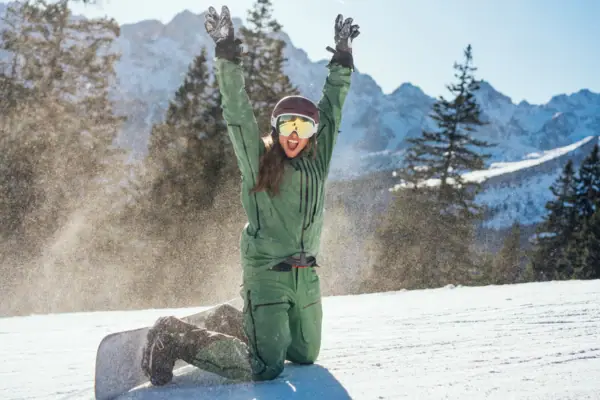A woman in winter clothing kneels in the snow with her hands raised.