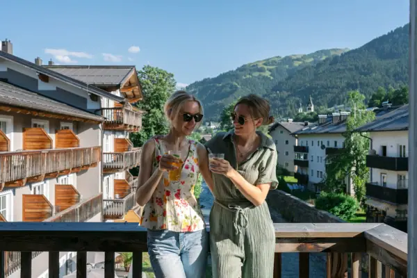 Two women stand on a balcony with a view of mountains and trees.