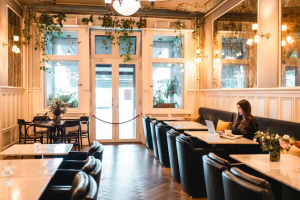 HENRI Hotel Berlin Salon breakfast area A young woman works on her laptop in the stylish hotel lobby of the HENRI Berlin, surrounded by classic wood panelling, marble tables and hanging green plants in the Belle Époque style.