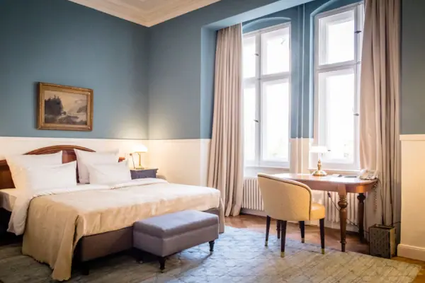 HENRI Hotel Berlin: Blue Design Room with Desk A stylish hotel room at the HENRI Berlin, featuring light blue walls, a double bed, an antique wooden desk and tall windows, all set within a classic historic building.