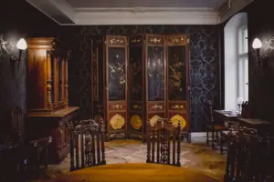 A magnificently designed salon at HENRI Berlin, featuring dark patterned wallpaper, an antique multi-panel screen with gold paintwork, and stately wooden furniture.