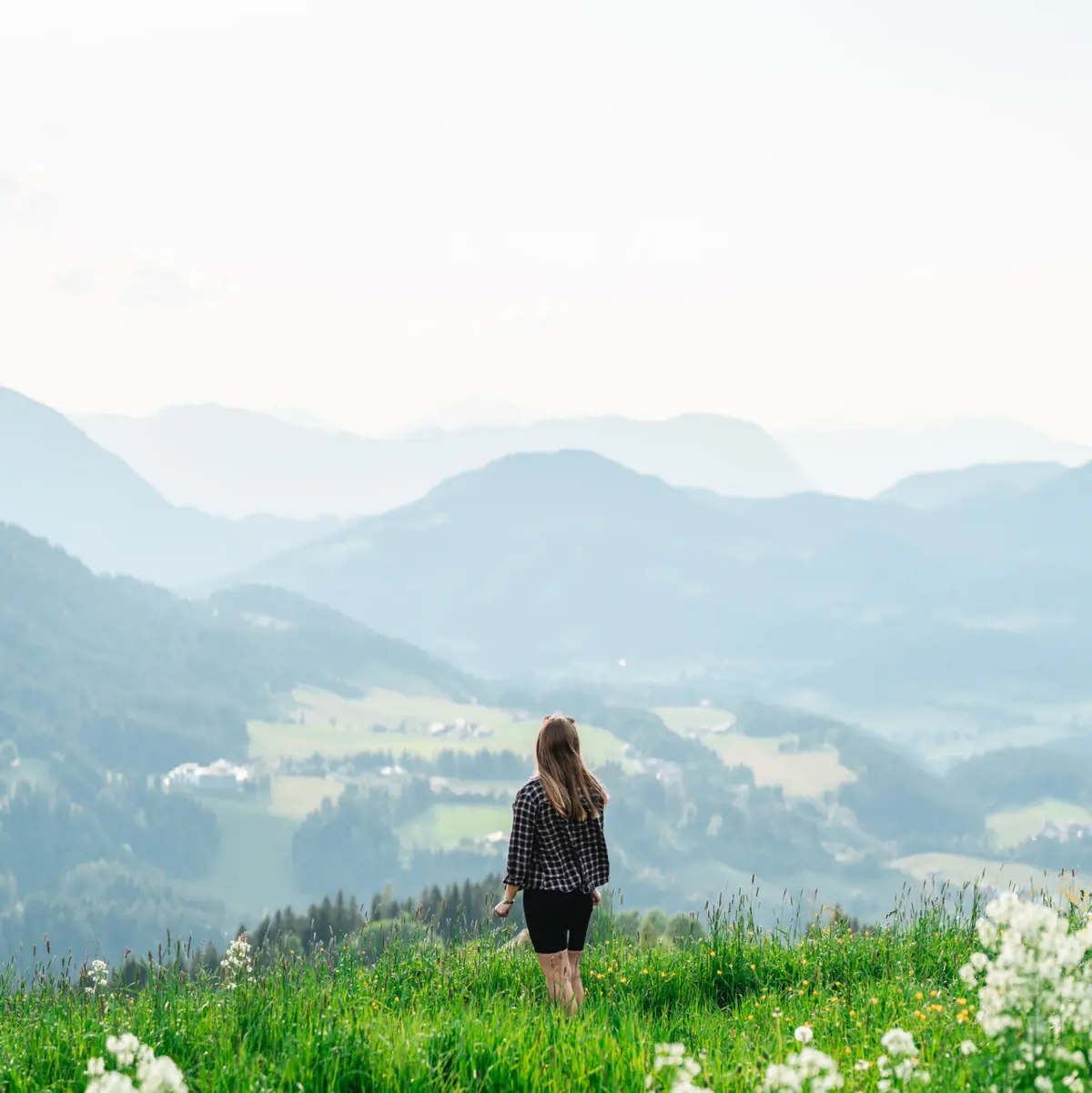 A woman stands on a grassy hill with mountains in the background.