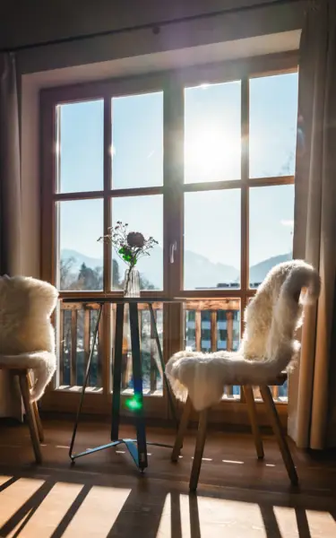 A cozy indoor scene with two wooden chairs draped in soft white sheepskin facing a sunlit window. A small round table with a glass vase holding flowers stands between the chairs, with sunlight streaming through the window.