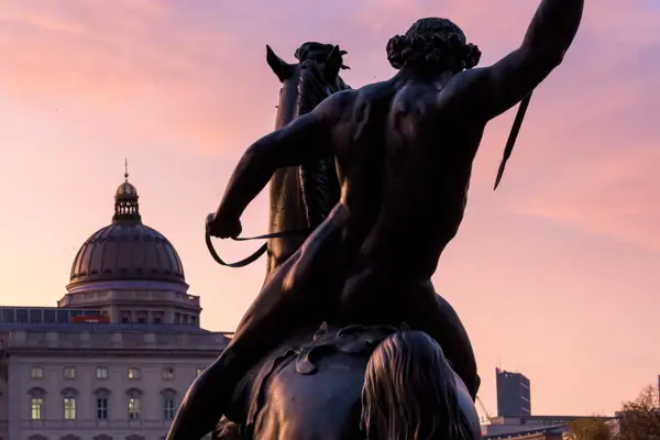 he dark silhouette of a monumental equestrian statue in Berlin against an atmospheric, pinkish-purple evening sky with a dome in the background.