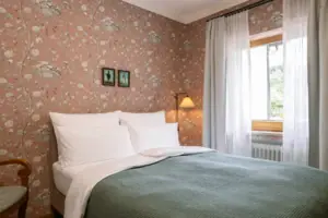 A bed with a green bedspread and a lamp against a floral wallpaper backdrop.
