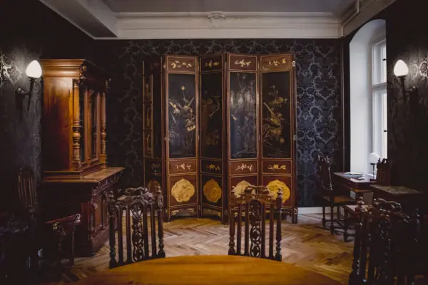 HENRI Hotel Berlin: Salon interior with a folding screen A magnificently designed salon at HENRI Berlin, featuring dark patterned wallpaper, an antique multi-panel screen with gold paintwork, and stately wooden furniture.