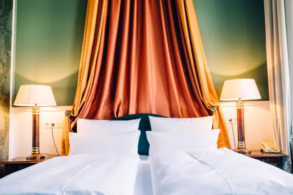 HENRI Hotel Berlin Room Details: Canopy A close-up of a stately hotel bed at the HENRI Berlin, featuring a magnificent orange-and-gold canopy against a green wall and classic bedside lamps.