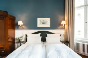 Front view of a stylish double bed in the HENRI Berlin with deep blue wall, classic wooden wardrobe, symmetrical wall lights and a framed architectural print.