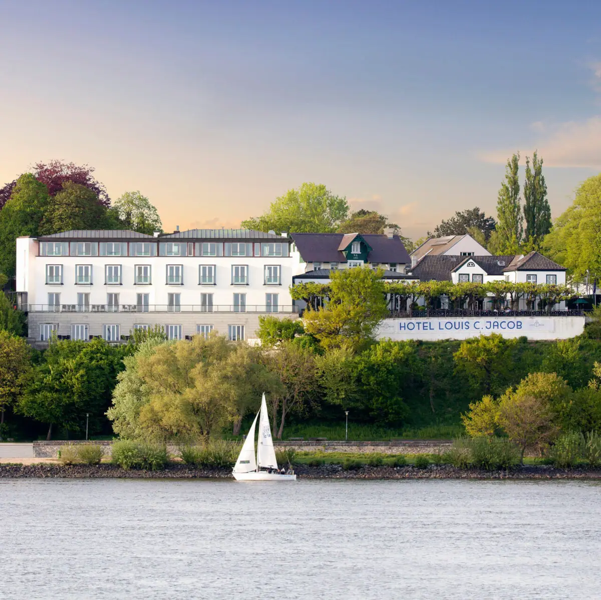 A white building with a sailing boat in front of a body of water.