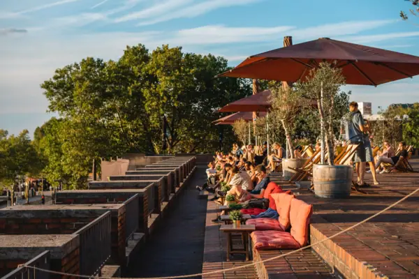 A group of people sitting on a patio under umbrellas with trees and plants nearby.
