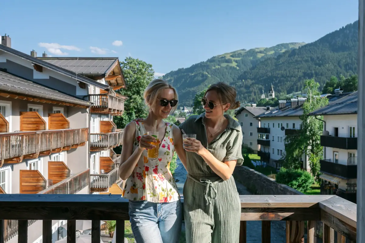 Two women stand on a balcony with a view of mountains and trees.
