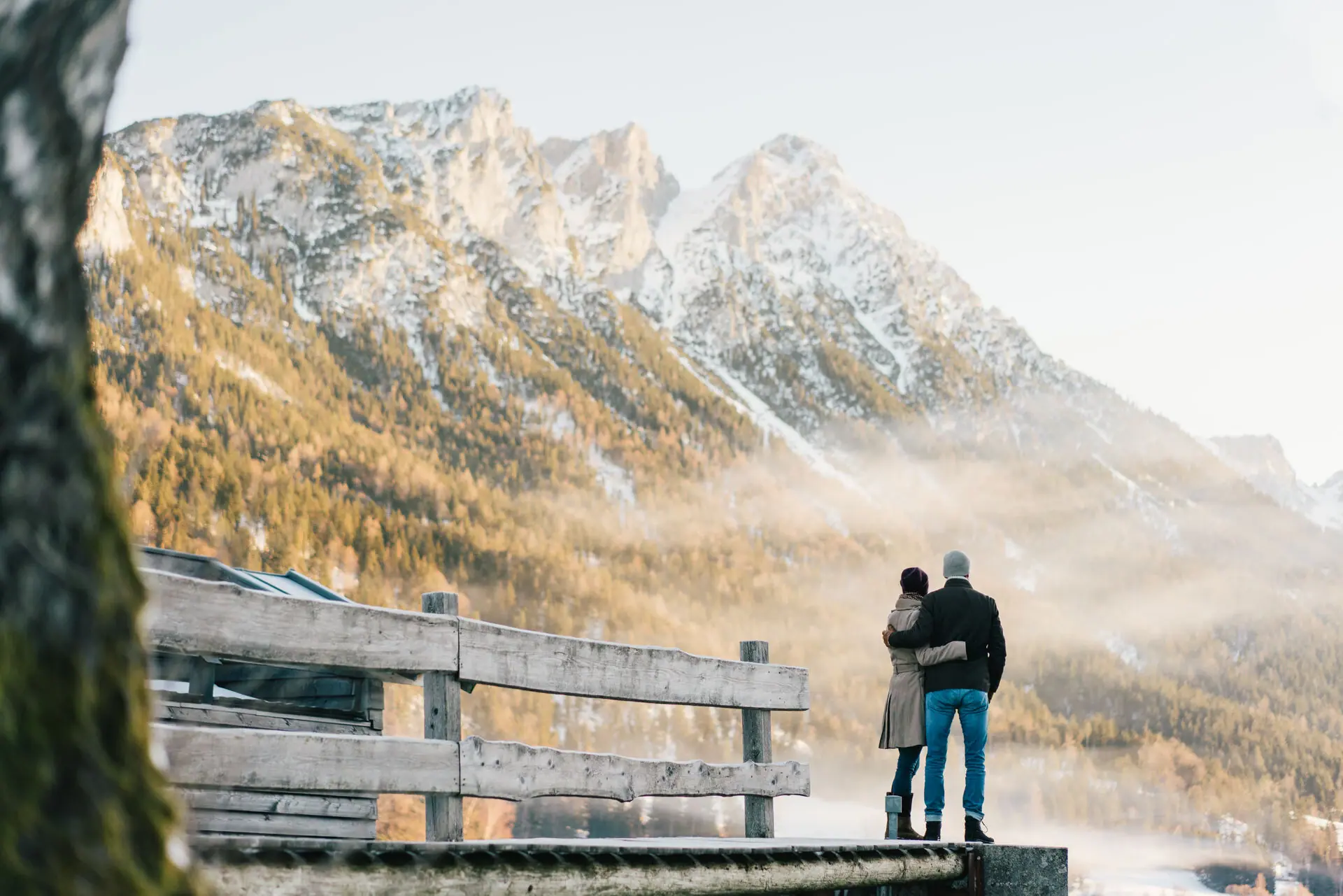 A man and a woman are standing on a jetty overlooking a mountain range.