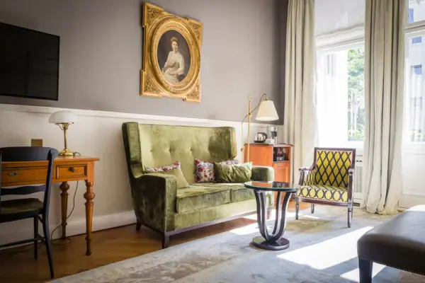 Klassisches Hotelzimmer im Gründerzeit-Stil mit einem grünen Samtsofa, antikem Schreibtisch und herrschaftlichem Porträtgemälde an der Wand.
