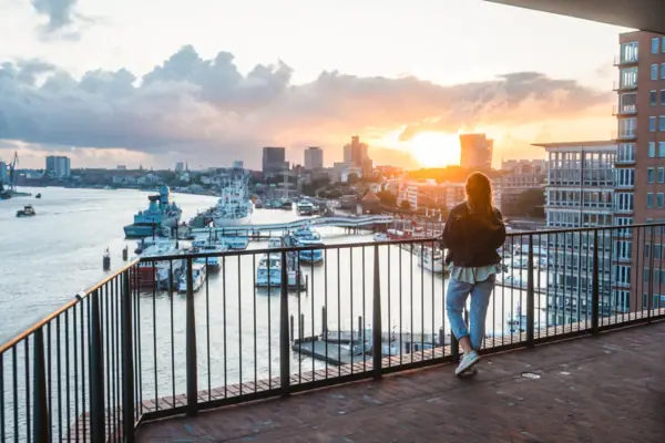 A woman stands on a balcony and looks out over a body of water with boats in the background.