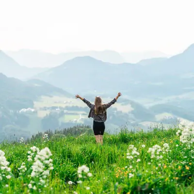 A woman stands in a field of flowers with her arms raised.