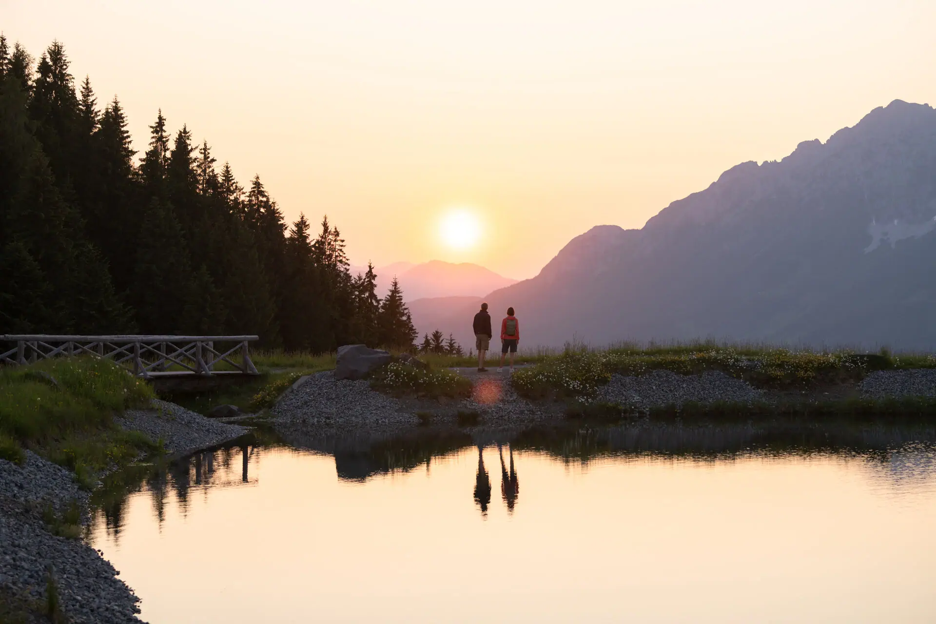Two people walk along the rocky shore of a lake, with trees and mountains in the background.