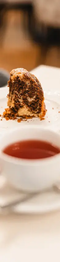 Close-up of a slice of marble cake served on a white plate alongside a cup of tea in a white porcelain cup held by a hand. 