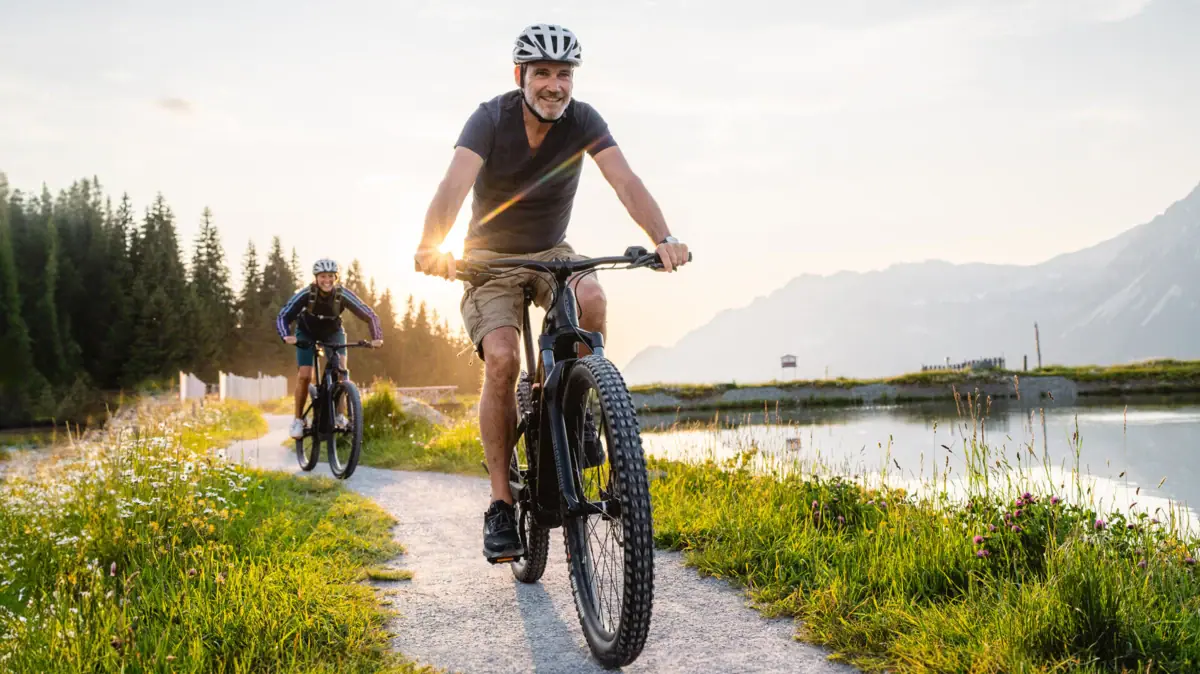 A man is cycling along a path; a lake and mountains can be seen in the background.