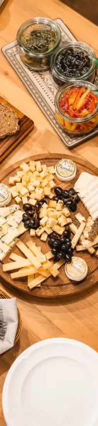 Wooden table with a cheese platter featuring a variety of cheesesand a stack of white plates with cutlery nearby.