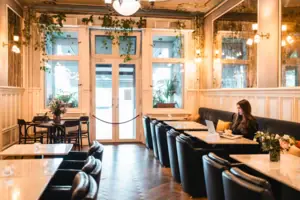 A young woman works on her laptop in the stylish hotel lobby of the HENRI Berlin, surrounded by classic wood panelling, marble tables and hanging green plants in the Belle Époque style.